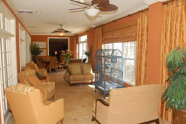 A warmly decorated living room area with wicker and cushioned chairs, a loveseat, a large birdcage, and tropical-themed ceiling fans. The room has large windows with bamboo blinds and patterned curtains, and there are plants and a dining area visible in the background.