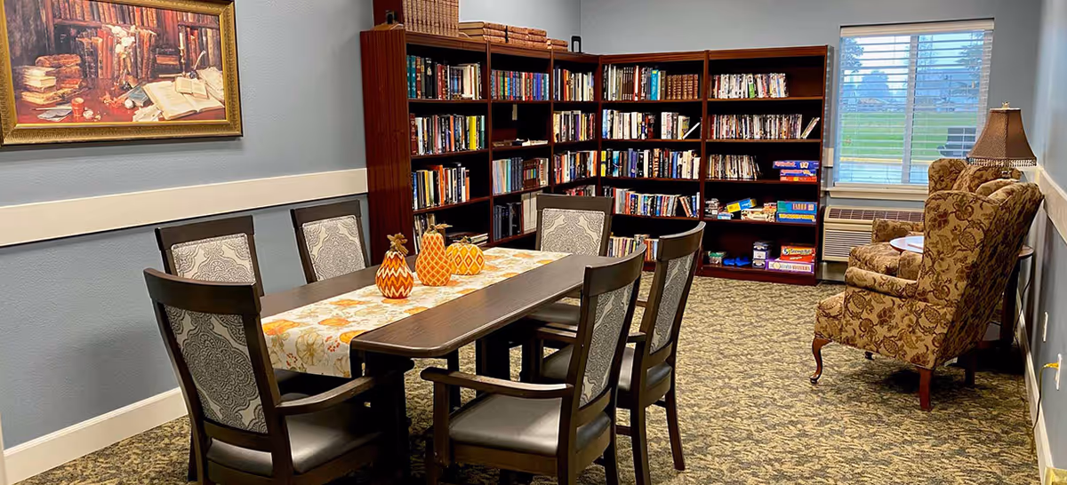 A cozy room with a wooden table and six chairs arranged around it, decorated with a floral table runner and three decorative pumpkins. Behind the table, there are bookshelves filled with books and board games. To the right, there are two upholstered armchairs next to a window with blinds and a small side table with a lamp. The room has carpeted flooring and light blue walls with a framed painting hanging on the left wall.
