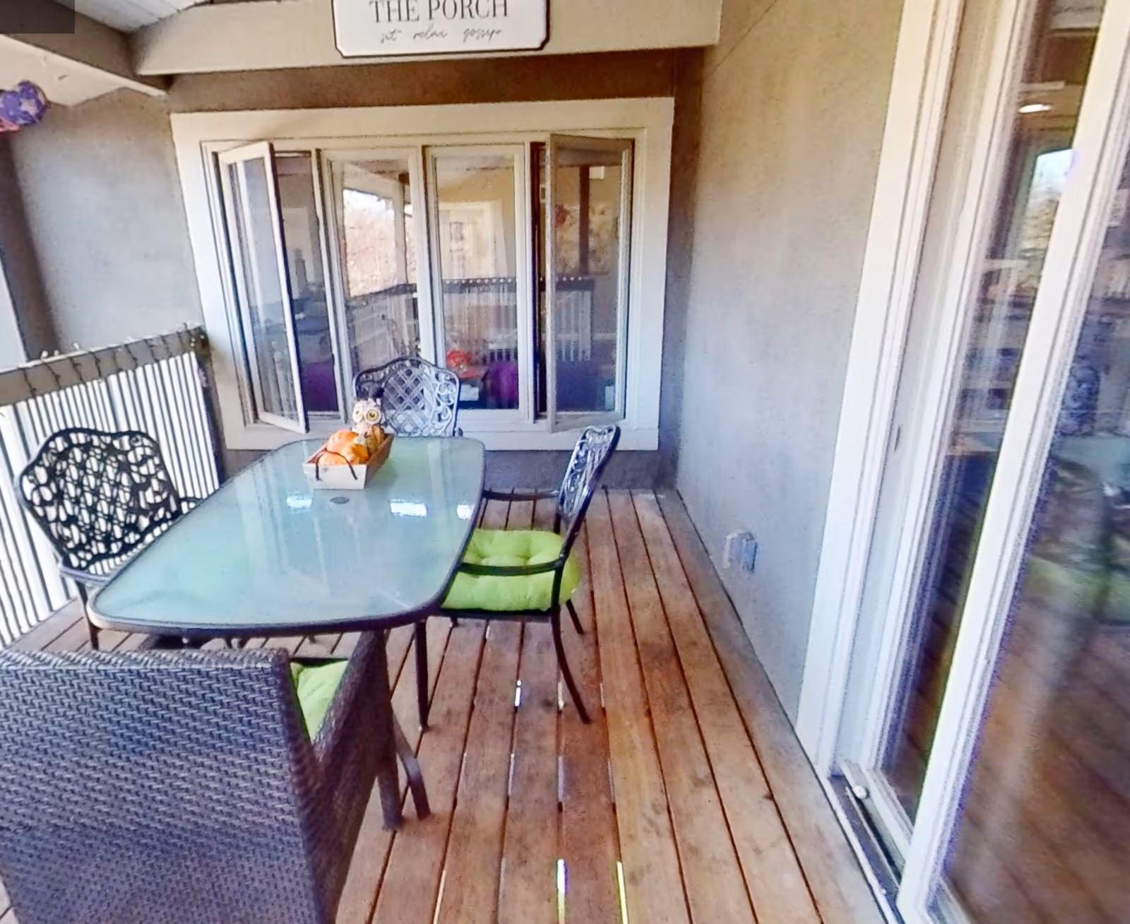 A covered porch area with wooden flooring featuring a glass-top table surrounded by four chairs with green cushions. The porch has a railing on one side and large windows and a sliding glass door on the other side. A decorative sign above the windows reads 'THE PORCH sit relax gossip'.