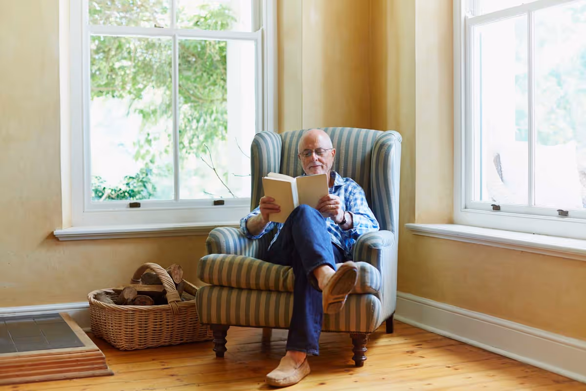 An elderly man wearing glasses and a blue checkered shirt is sitting comfortably in a striped armchair by two large windows, reading a book. There is a basket filled with firewood on the wooden floor next to the chair.