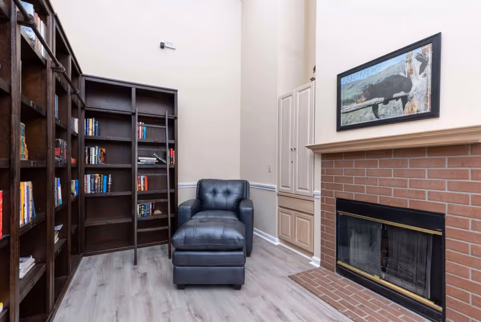A cozy reading nook featuring a dark leather armchair with an ottoman, positioned next to tall dark wood bookshelves filled with books. The room has light-colored walls and flooring, a brick fireplace with a black and gold screen, and a framed picture of a bear labeled 'Lake Tahoe' hanging above the fireplace.
