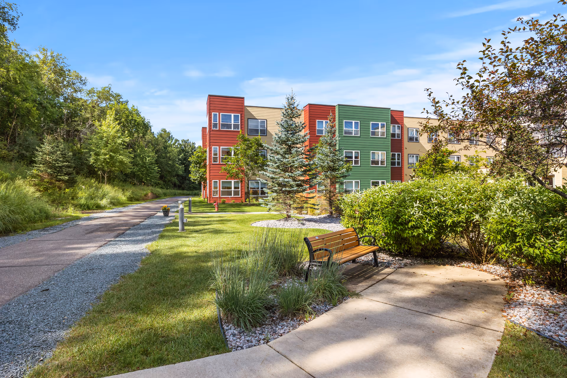 Outdoor view of Woodland Hill facility showing a colorful three-story building with red, green, and beige sections. In the foreground, there is a paved walkway, a wooden bench, green bushes, and trees under a clear blue sky.