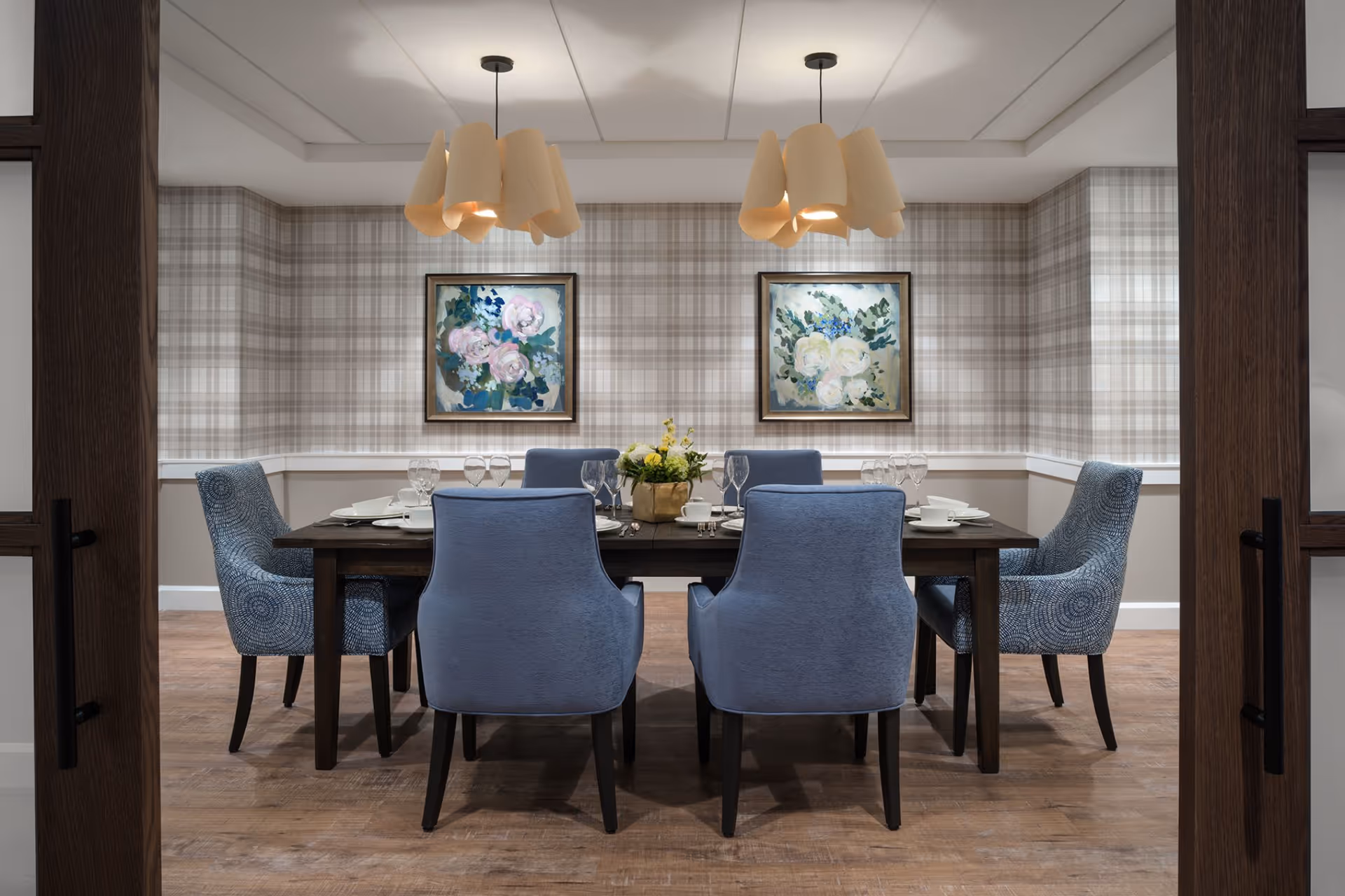 A dining room with a dark wooden table set for six people with blue upholstered chairs. The walls have a plaid wallpaper with two framed floral paintings. Two modern pendant lights hang from the ceiling above the table. The floor is wooden, and the room is viewed through open double doors with dark wood frames.