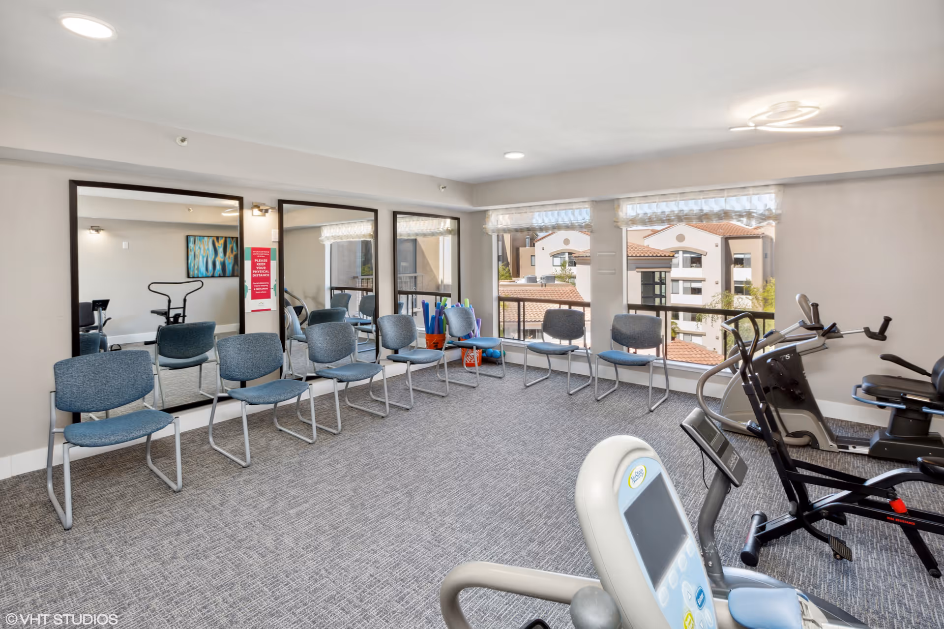 A fitness room with exercise equipment including stationary bikes and chairs arranged along the walls. Large mirrors and windows provide natural light and a view of nearby buildings with red-tiled roofs.