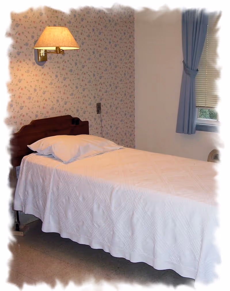 A simple single bed with a white quilt and pillow in a room with floral wallpaper on one wall and a window with blue curtains on the adjacent wall. A wall-mounted lamp is above the headboard.