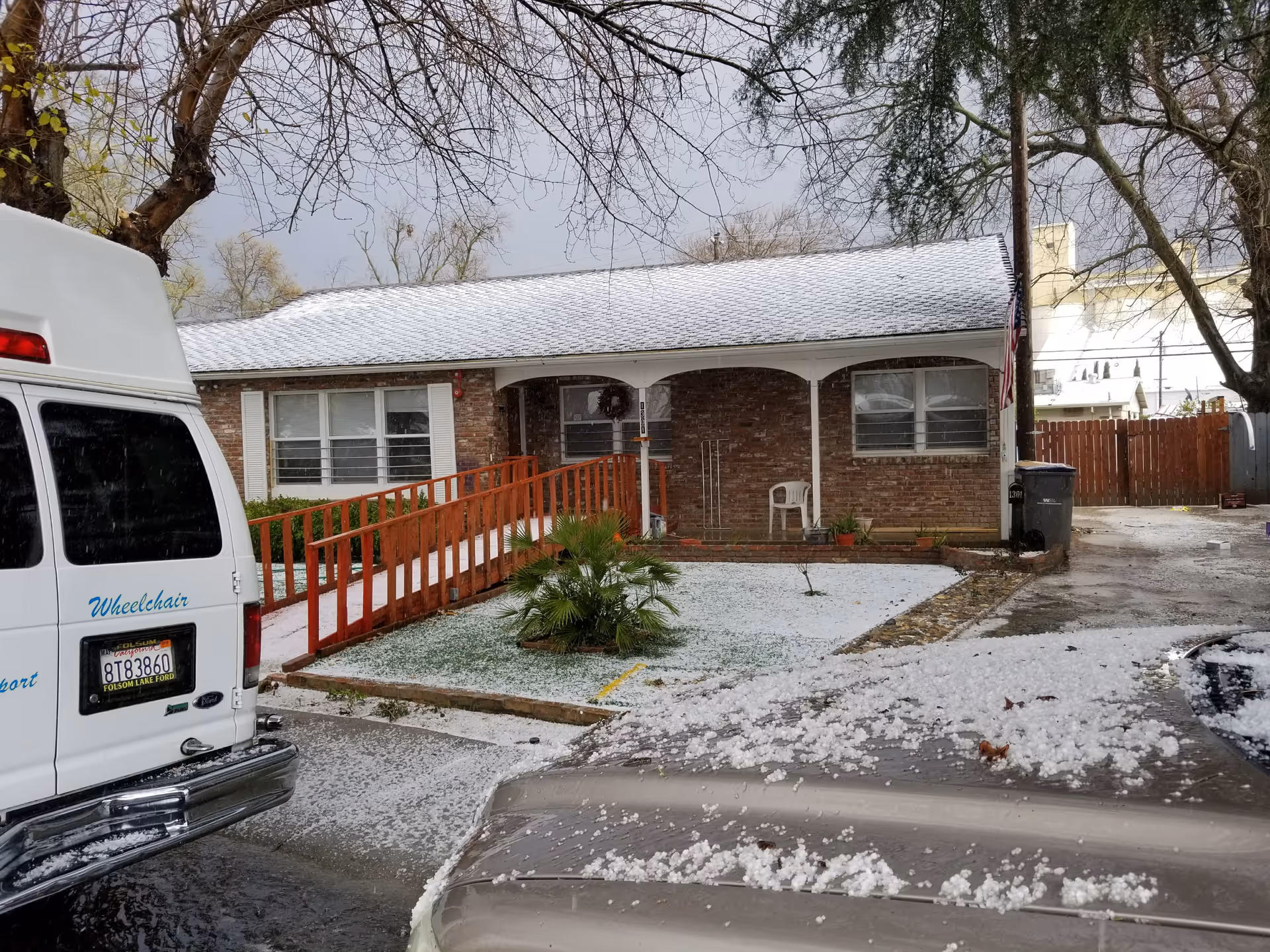 Exterior view of a single-story brick residential care facility with a snow-covered roof and ground. There is a wooden ramp leading to the entrance, a small palm plant in the front yard, and an American flag on a pole near the building. Two vehicles are partially visible in the foreground, one of which is a white van with the word 'Wheelchair' written on it.