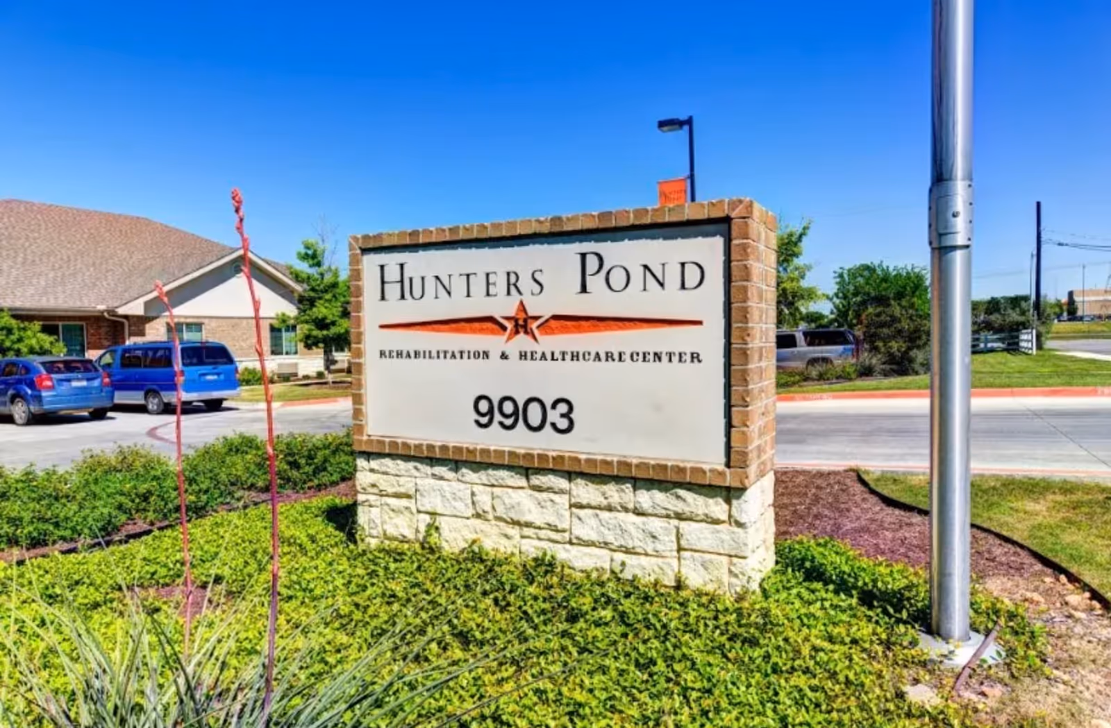 Outdoor view of a sign for Hunters Pond Rehabilitation and Healthcare Center with the address number 9903, surrounded by greenery and a clear blue sky in the background.