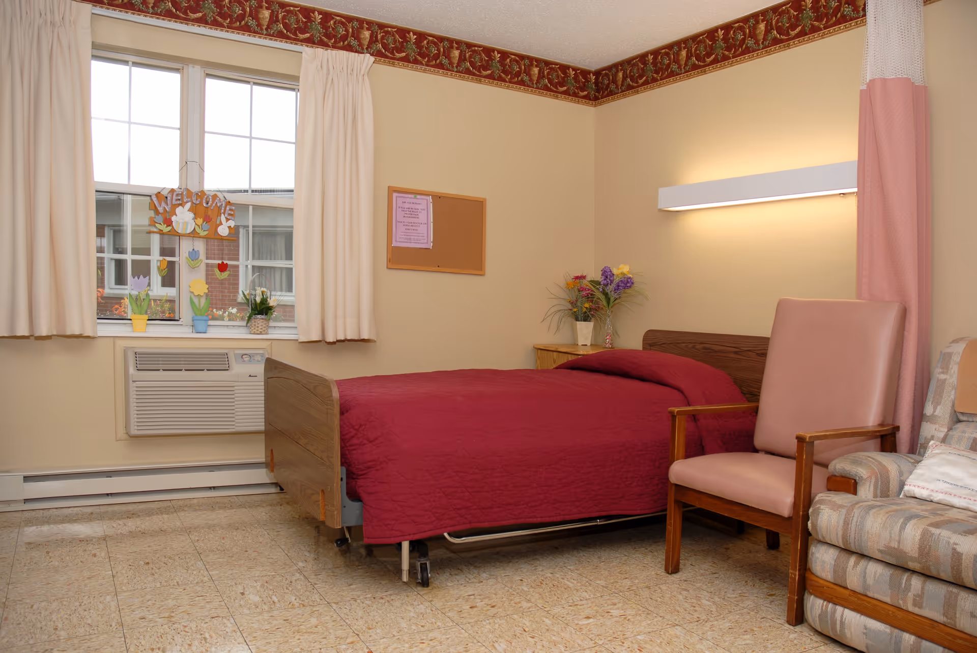 A tidy senior bedroom with a single bed dressed in a red quilt, a window with curtains and air conditioner, a nightstand with flowers, and a pair of chairs.