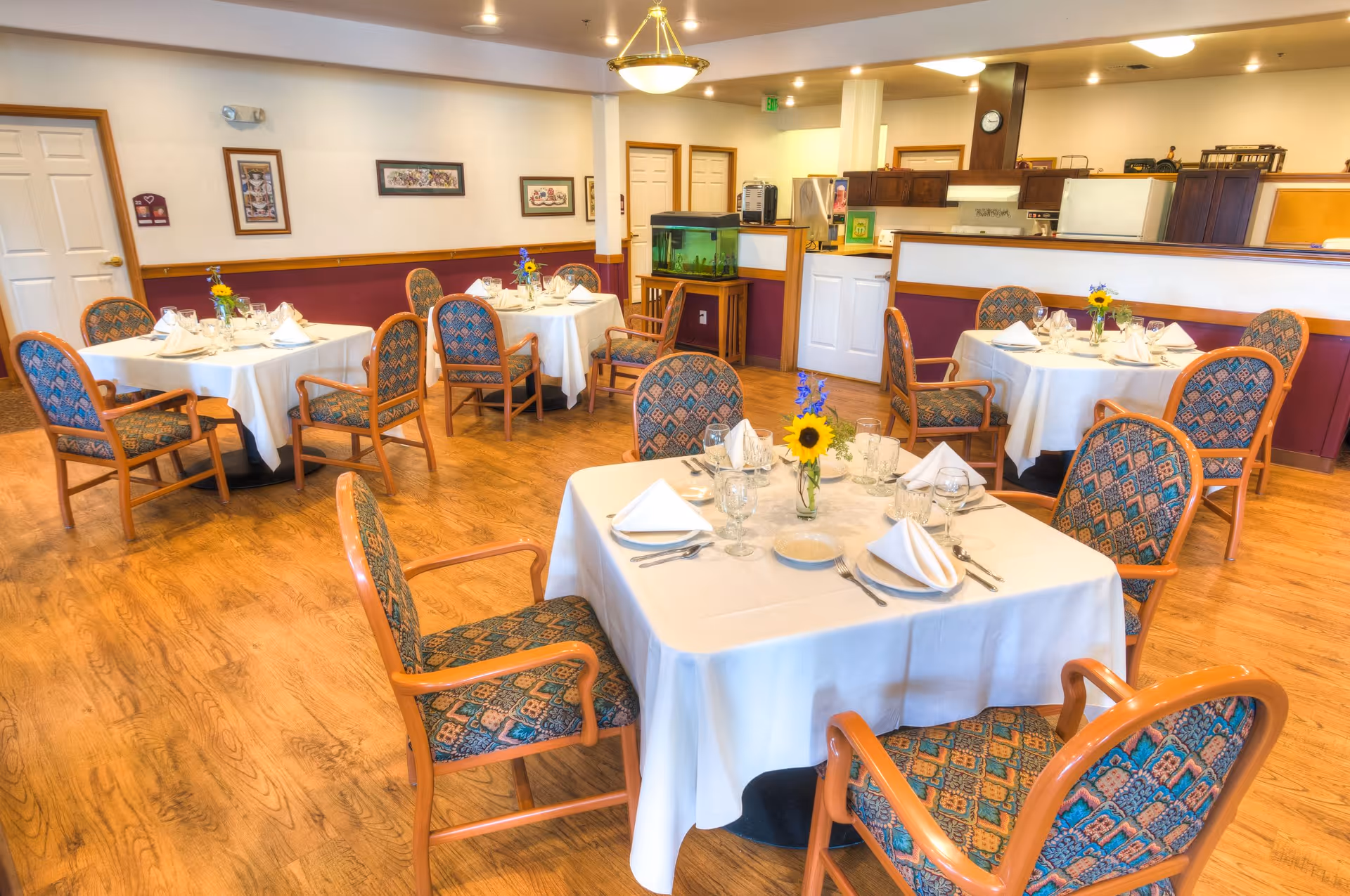 A dining room with several round tables covered with white tablecloths, each set with plates, glasses, napkins, and silverware. Each table has a small vase with a sunflower and other flowers. The chairs around the tables have wooden frames and patterned upholstery. The room has wooden flooring, framed pictures on the walls, and a fish tank near a counter that separates the dining area from a kitchen or service area in the background.