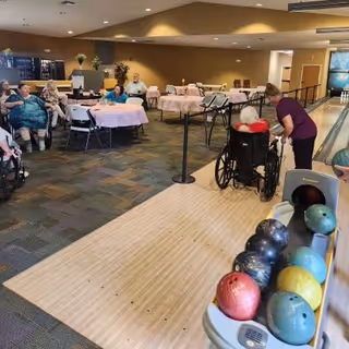 A group of elderly people sitting around tables covered with pink tablecloths in a large room. One elderly person in a wheelchair is being assisted by a caregiver near a bowling lane with several bowling balls visible in the foreground.