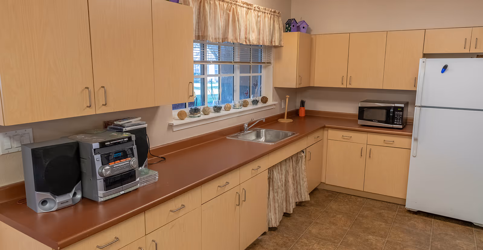 Communal kitchen with light wood cabinets, brown countertops, a sink under a window, microwave, refrigerator and a stereo on the counter.