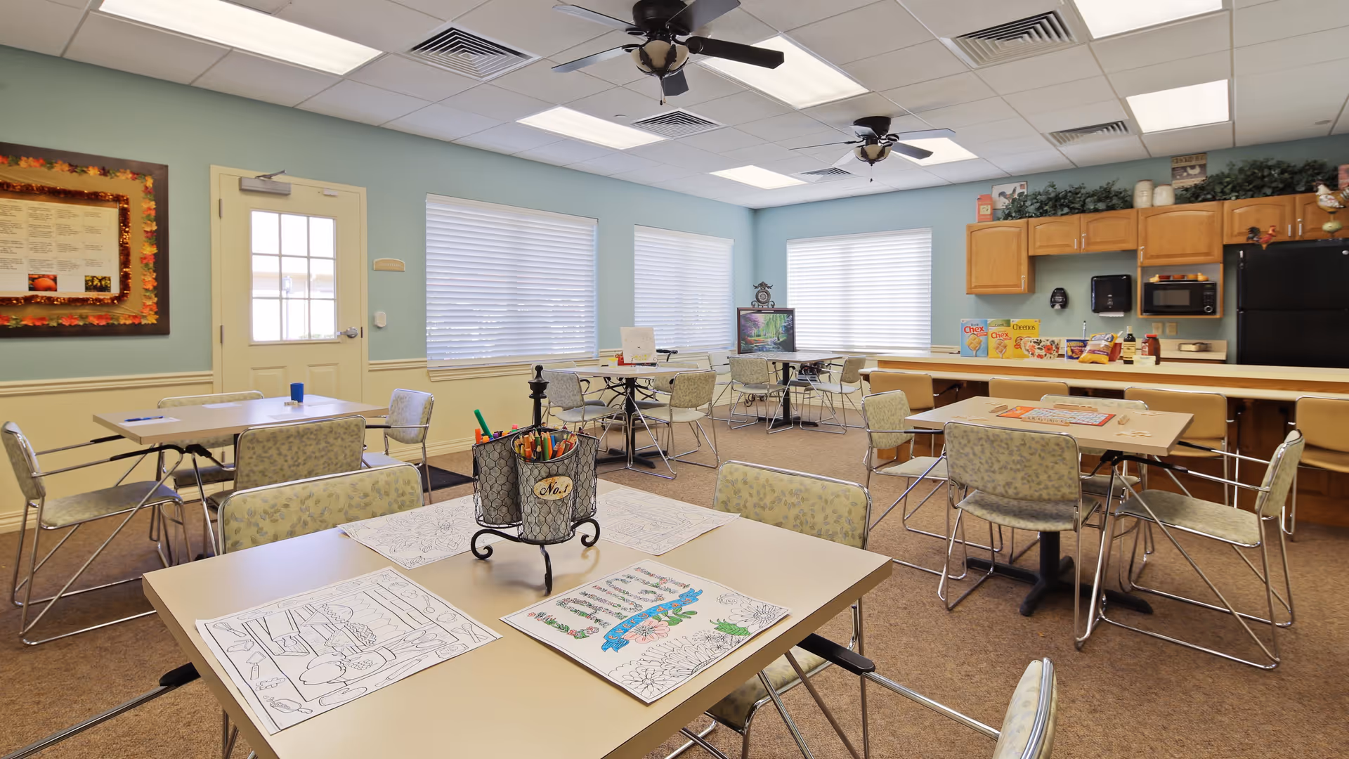 A bright activity room with several tables and chairs arranged for group activities. Coloring sheets and colored pencils are placed on the tables. The room has light blue walls, large windows with blinds, ceiling fans, and a kitchenette area with cabinets, a microwave, and a refrigerator. There is a TV on a stand in the corner and some cereal boxes on the counter.