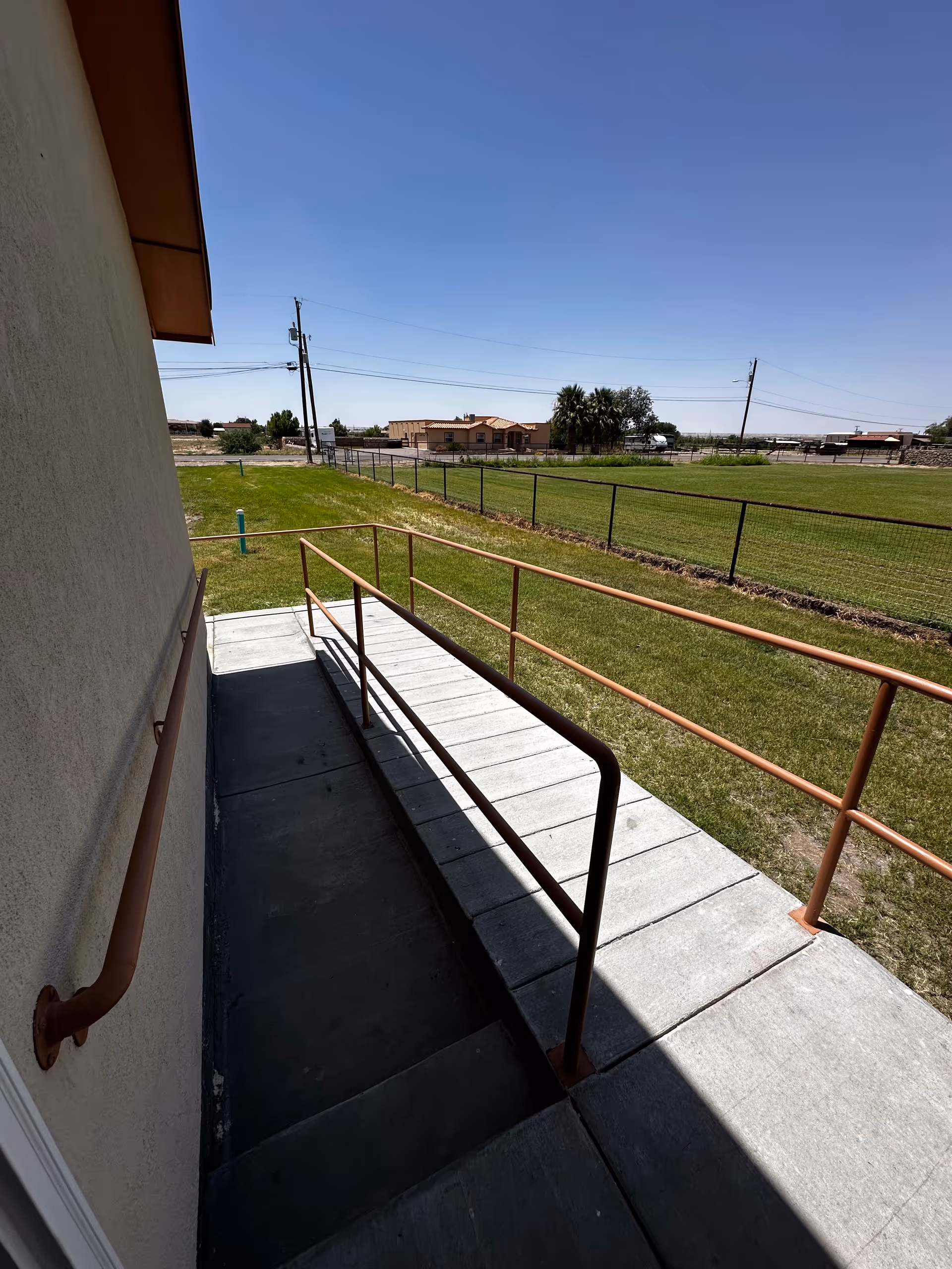 Exterior concrete ramp with metal handrails beside a building leading to a grassy yard and fence under a clear blue sky.