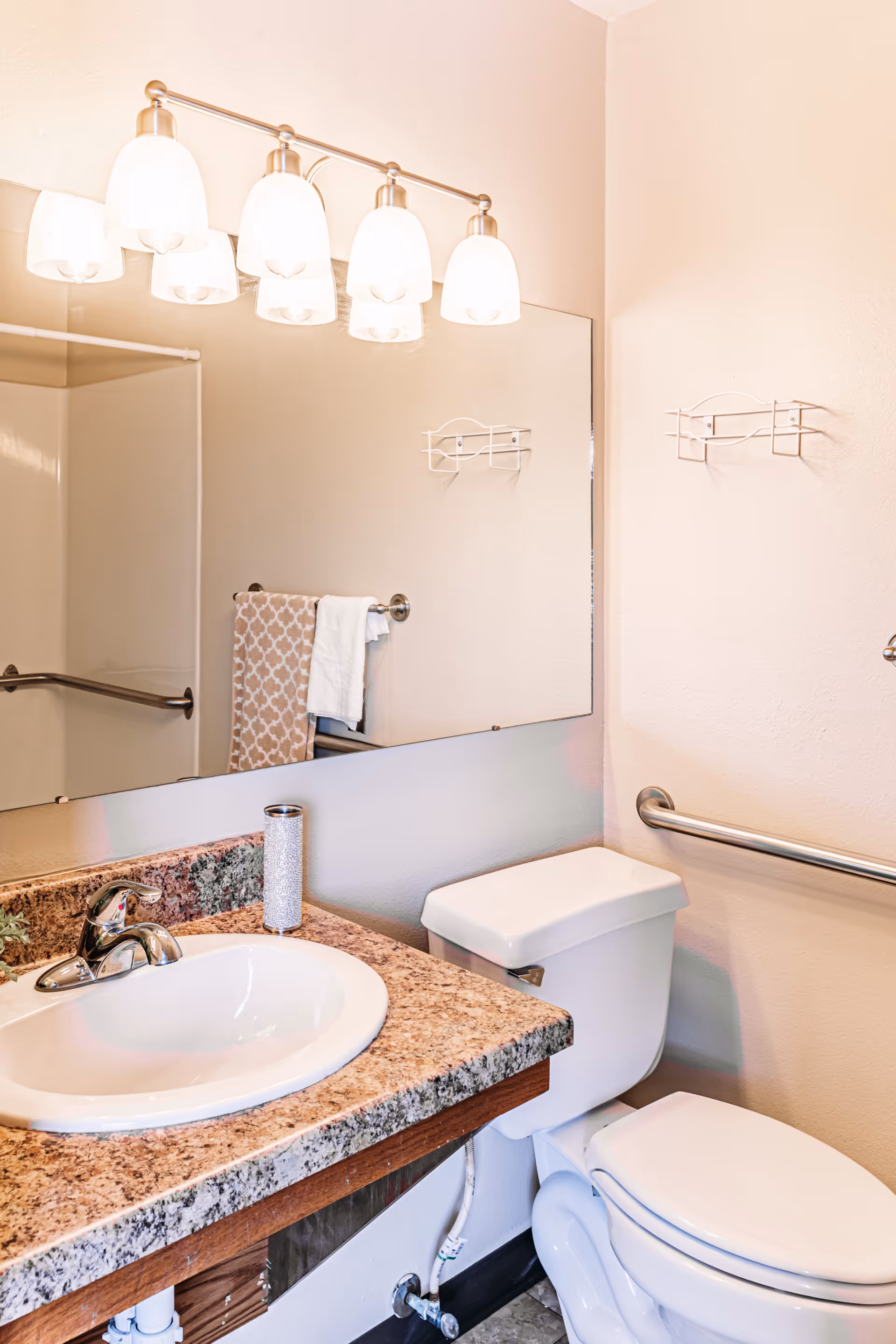 A clean bathroom featuring a white toilet and a sink with a granite countertop. Above the sink is a large mirror with a light fixture consisting of five frosted glass lamps. There is a towel rack with a patterned towel and a white towel, as well as a metal grab bar on the wall next to the toilet.