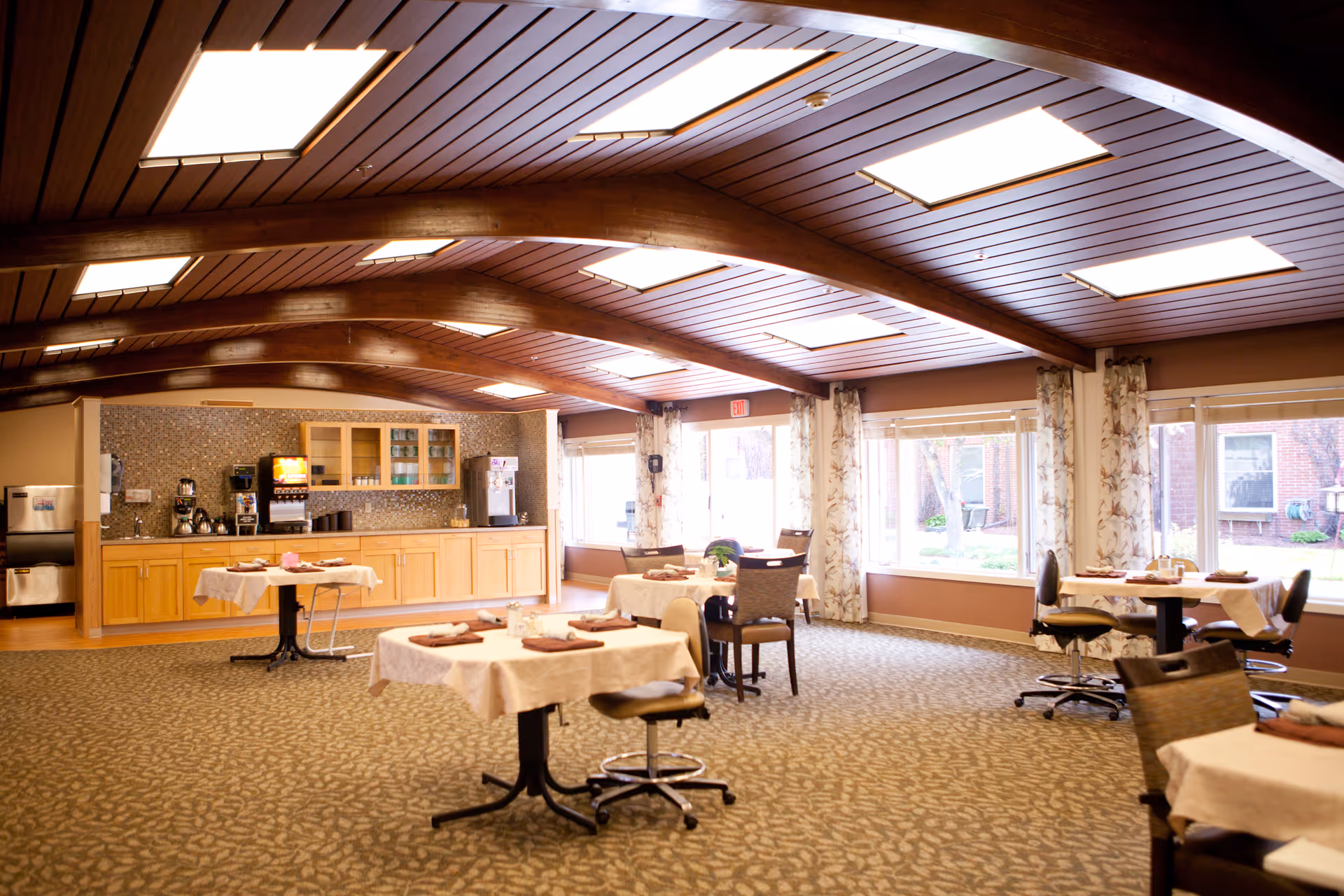 A spacious dining room with several tables set for meals, chairs, large windows, a wood-beamed ceiling, and a serving counter with coffee machines.