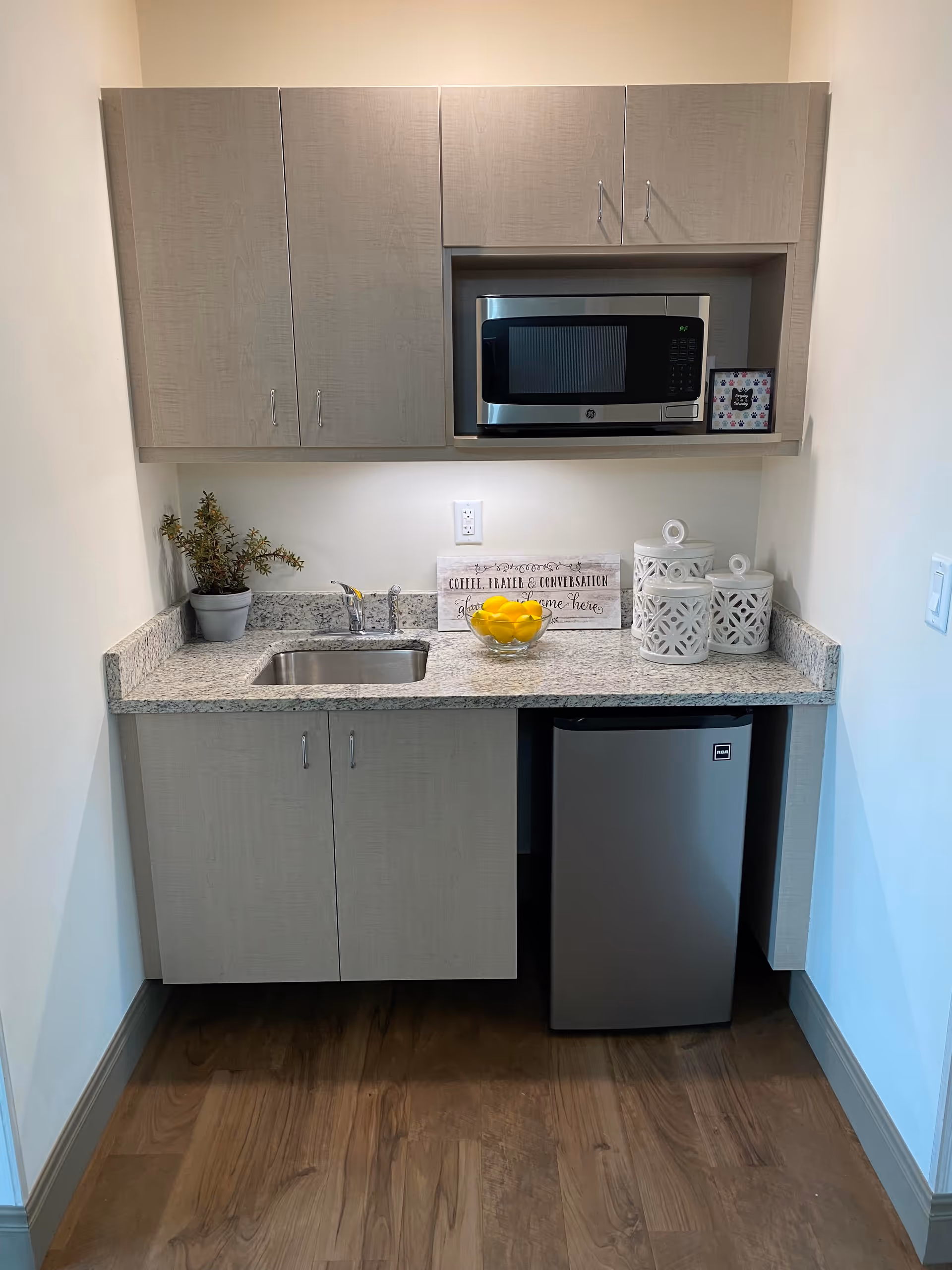 Small kitchenette with light wood cabinets, a granite countertop, a stainless steel sink, a microwave, a mini refrigerator, a bowl of lemons, decorative white canisters, a small potted plant, and a sign that reads 'Coffee, Treats & Conversation, Gather some here'.
