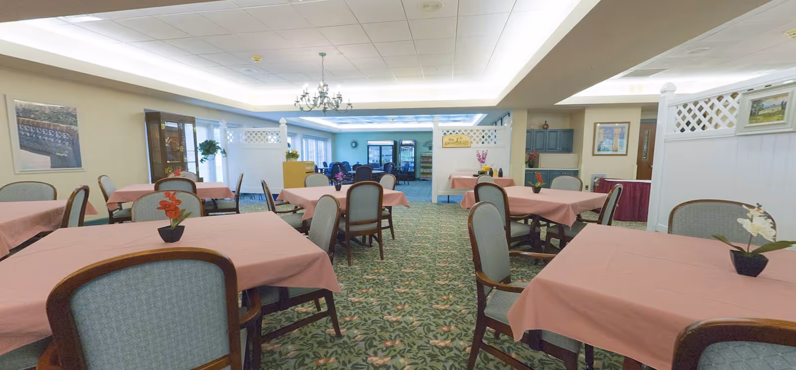 A dining room with multiple tables covered in pink tablecloths, each with a small flower arrangement. The room has patterned carpet, light blue cushioned chairs with wooden frames, and a chandelier hanging from the ceiling. In the background, there is a section with refrigerators and additional seating.