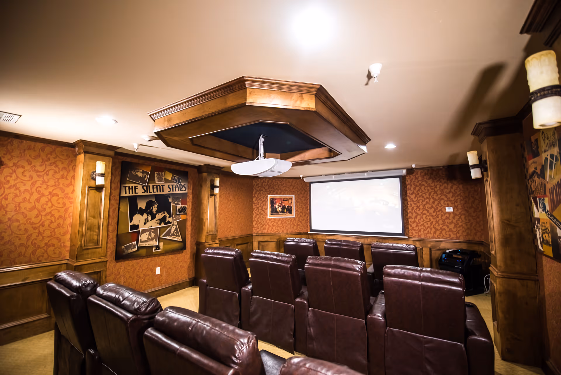 A cozy home theater room with two rows of brown leather recliner chairs facing a large projector screen. The walls are decorated with vintage movie posters and have warm, patterned wallpaper with wooden paneling. The ceiling features a decorative wooden structure with a ceiling fan.