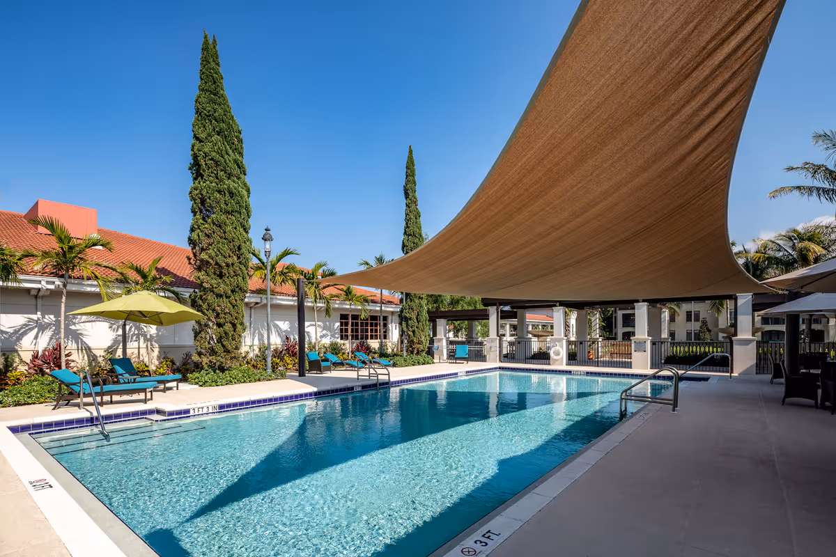 Outdoor swimming pool area with clear blue water, surrounded by lounge chairs with green umbrellas, tall cypress trees, and a large beige shade sail overhead. The pool area is adjacent to a building with a red-tiled roof and palm trees under a clear blue sky.