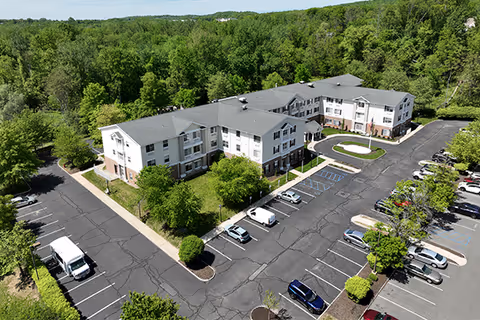 Aerial view of a three-story senior living building with a surrounding parking lot and wooded backdrop.
