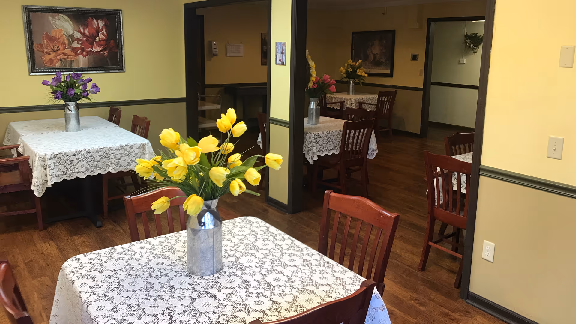 Interior view of a dining room in Blue Hills Rest Home with several wooden tables covered with white lace tablecloths. Each table has a metal vase with colorful artificial flowers, including yellow tulips and purple flowers. The walls are painted yellow with dark trim, and there is a framed floral painting on one wall. The floor is wooden.