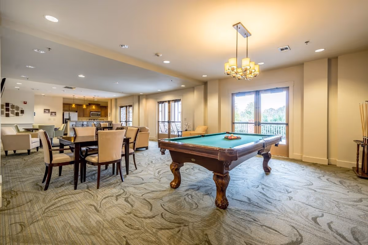 A spacious common area in a senior living facility featuring a pool table with balls arranged for a game, several dining tables with chairs, comfortable armchairs, and a kitchen area in the background. Large windows and glass doors allow natural light to fill the room.