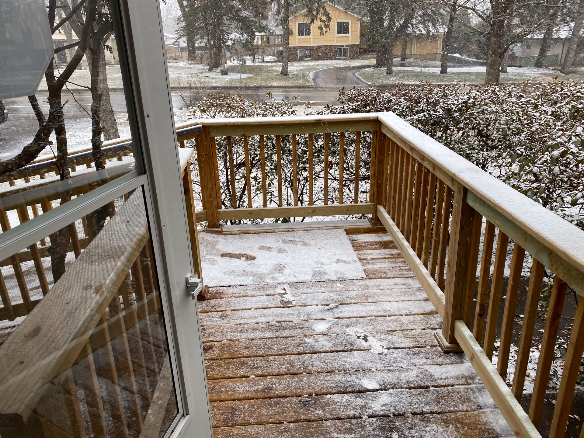Snow-dusted wooden porch with railings overlooking a street and houses.