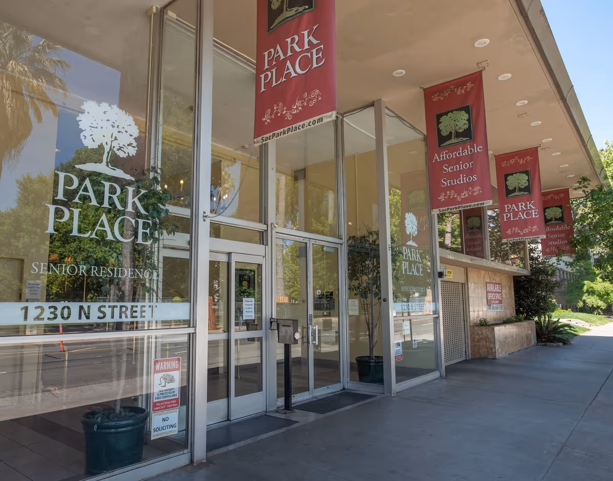 Glass-front entrance to Park Place senior residence with red banners, potted plants, and the address 1230 N Street.