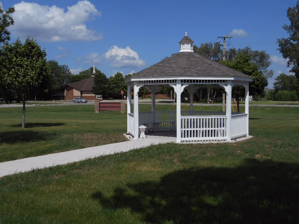 White octagonal gazebo on a grassy lawn with a sidewalk leading to it and trees and buildings in the background.