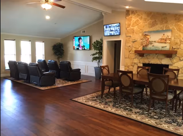 A spacious living room area with wooden flooring, featuring a row of black recliner chairs facing a wall-mounted TV. There is a dining table with six chairs on a patterned rug near a stone fireplace with a painting above it. Two potted plants are placed near the walls, and a smaller screen is mounted above a doorway.