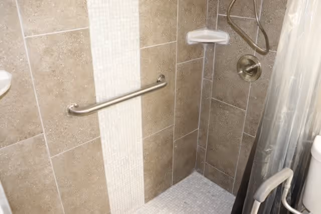 A tiled shower area with beige and white tiles, a metal grab bar on the wall, a soap holder, a handheld showerhead, and a shower curtain partially visible. Part of a toilet with a grab bar is also visible on the right side.