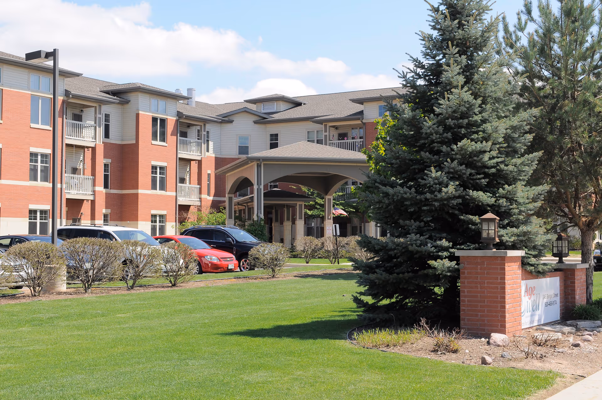 Exterior view of a multi-story senior living facility with red brick and beige siding. There is a covered entrance, several parked cars, trimmed bushes, green lawn, and tall evergreen trees near a brick sign.