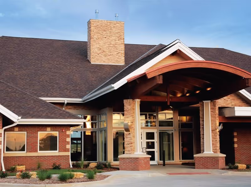 Front exterior view of Silvercrest at Woodlands Creek facility showing a brick building with large windows, a prominent stone chimney, and a covered entrance with wooden beams under a clear blue sky.