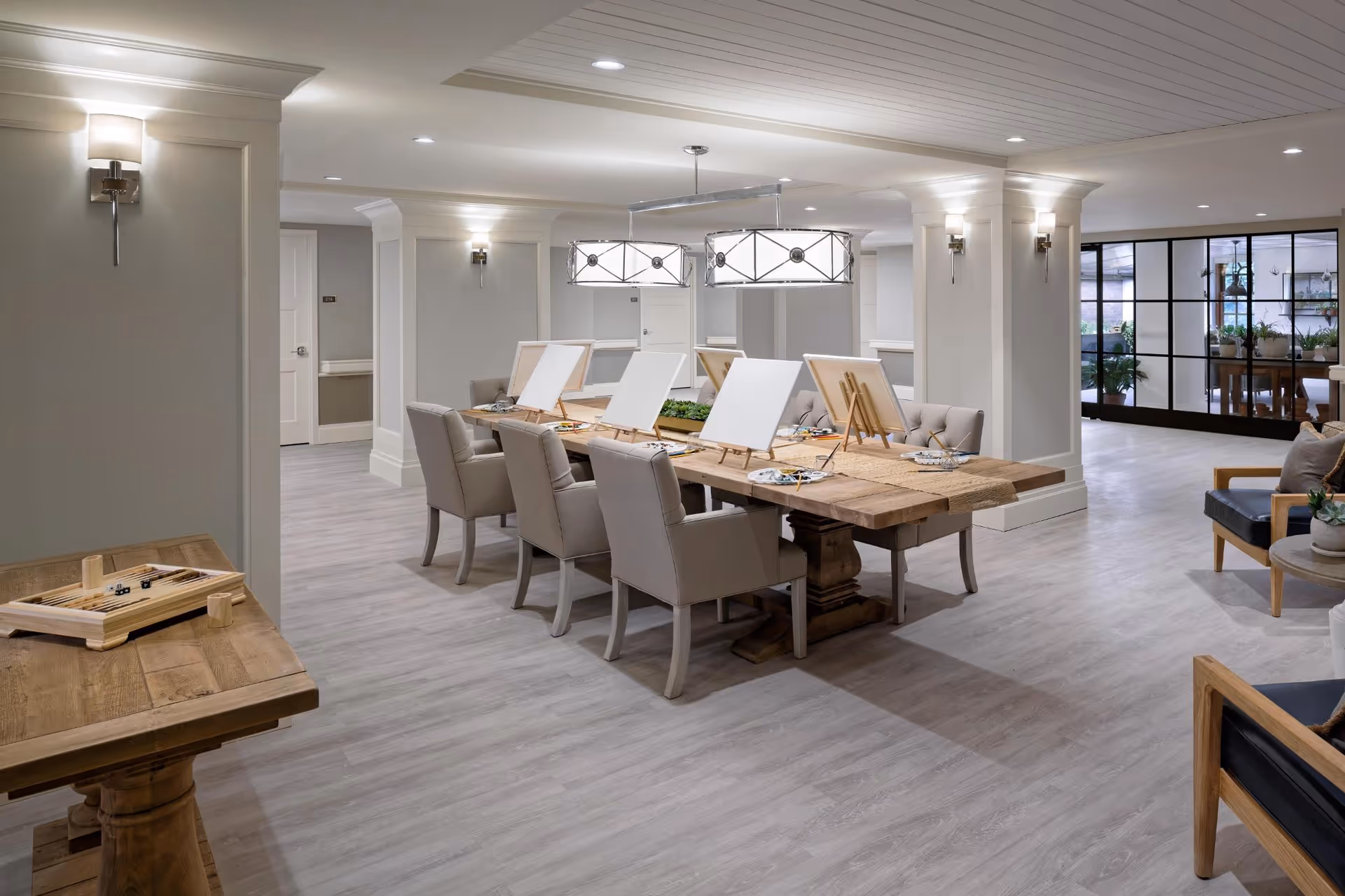 A bright, spacious activity room in a senior living facility featuring a large wooden table with six chairs, each with a blank canvas on an easel and painting supplies. The room has light gray walls, wood flooring, modern wall sconces, and two large pendant lights hanging above the table. In the foreground, there is a smaller wooden table with a board game. The background shows a seating area with armchairs and a glass partition with plants visible behind it.