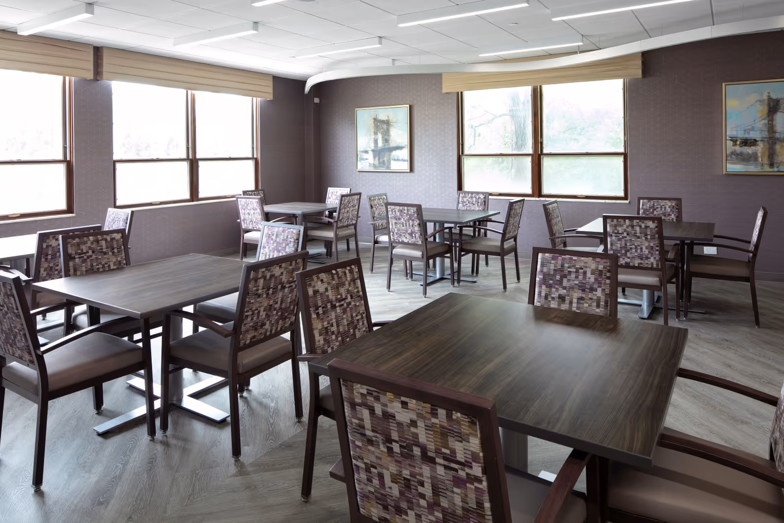 Bright empty dining room with multiple dark wood square tables and patterned upholstered chairs beside large windows.