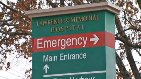 Green hospital sign reads Lawrence & Memorial Hospital Emergency Main Entrance with trees behind
