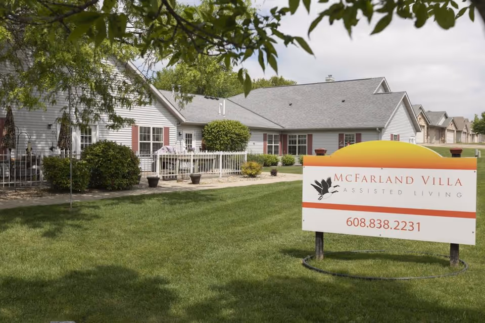 Exterior view of McFarland Villa Assisted Living facility showing a single-story building with white siding and red shutters, surrounded by green grass and trees. A sign in the foreground displays the facility name and phone number.
