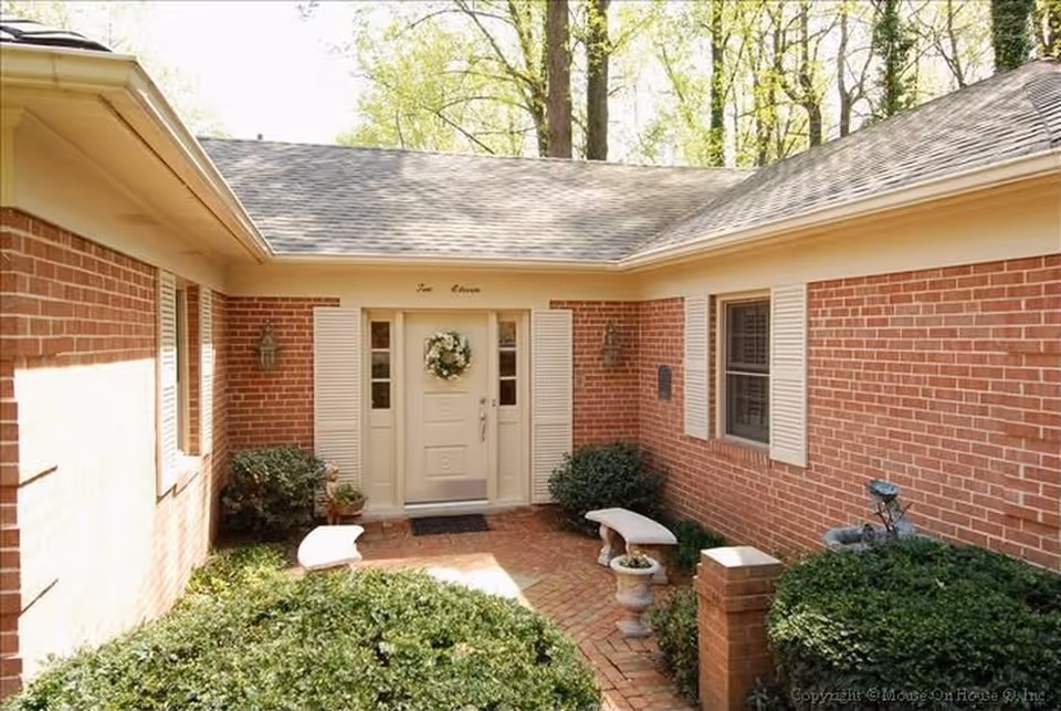 Entrance to a brick building with a white door decorated with a wreath, flanked by white shutters and outdoor wall lanterns. There are two white benches and green bushes along a brick-paved walkway leading to the door.