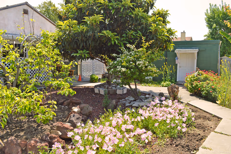 A garden area with various plants and flowers, including pink blooms in the foreground, small trees, and shrubs. There is a concrete pathway leading to a green building with a white door and a small American flag hanging beside it. A house with a porch and lattice railing is visible on the left side.