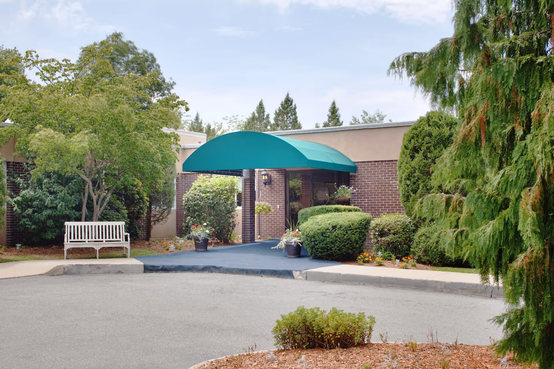 Entrance of a building with a green awning over the doorway, surrounded by bushes, trees, and flower pots. A white bench is placed on the left side near the entrance, and the sky is partly cloudy.