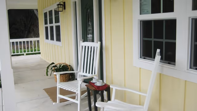 A porch area with two white rocking chairs and a small table between them holding two white cups and saucers. The porch has a yellow exterior wall with white trim around windows and a green door. A potted plant in a wicker basket is placed near the door.