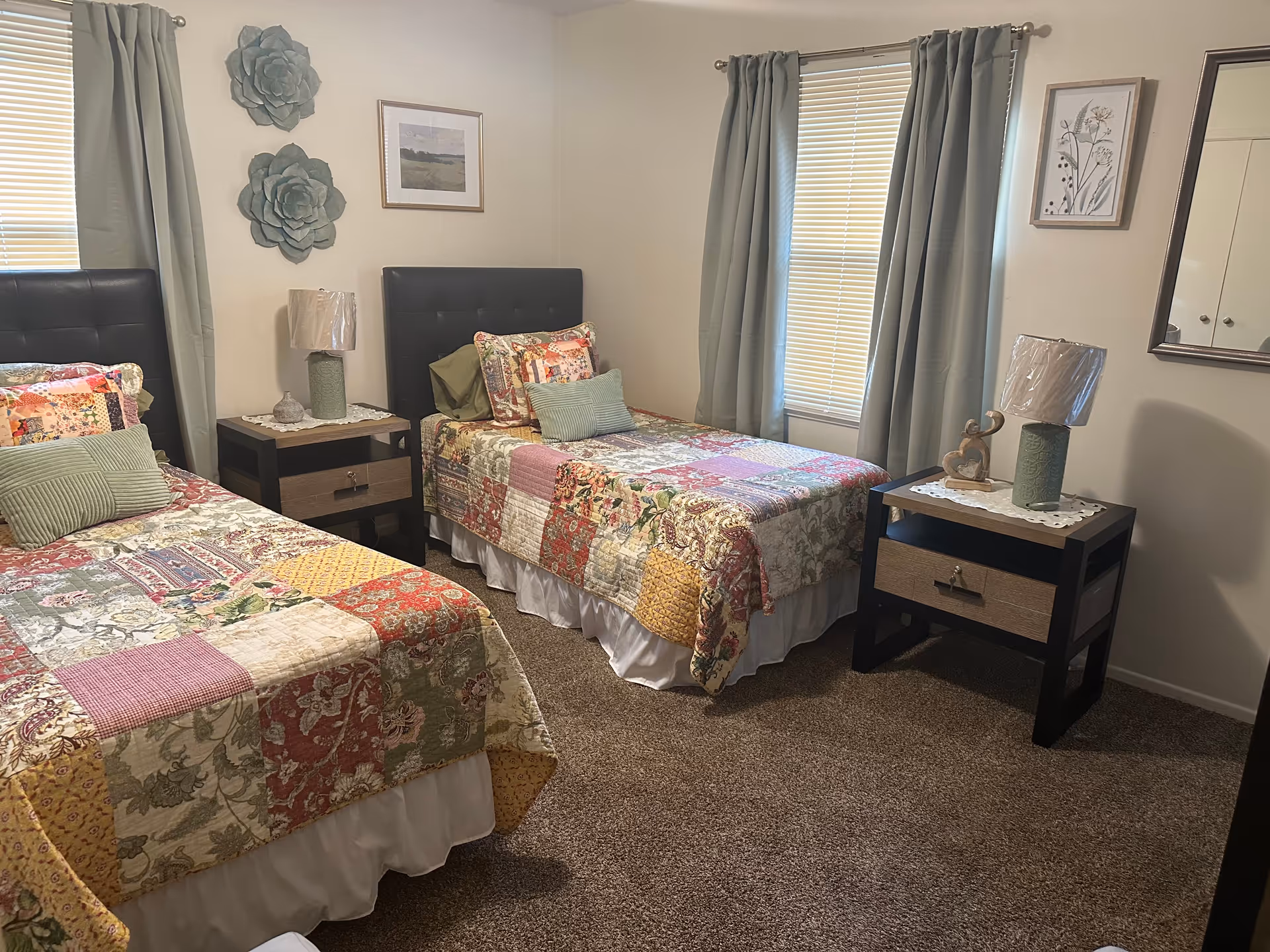 A bedroom with two twin beds featuring colorful patchwork quilts and green pillows. Each bed has a black tufted headboard. Between the beds are two matching nightstands with lamps, one with a green ceramic base and the other with a similar style lamp wrapped in plastic. The room has beige walls, two windows with closed blinds and green curtains, and decorative wall art including two large green flower sculptures and framed pictures. The floor is carpeted in brown.