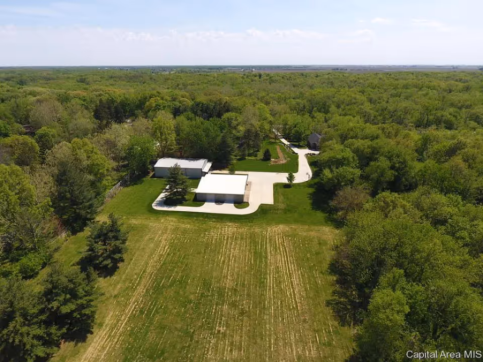 Aerial view of a house with outbuildings, driveway, and large lawns surrounded by dense trees.