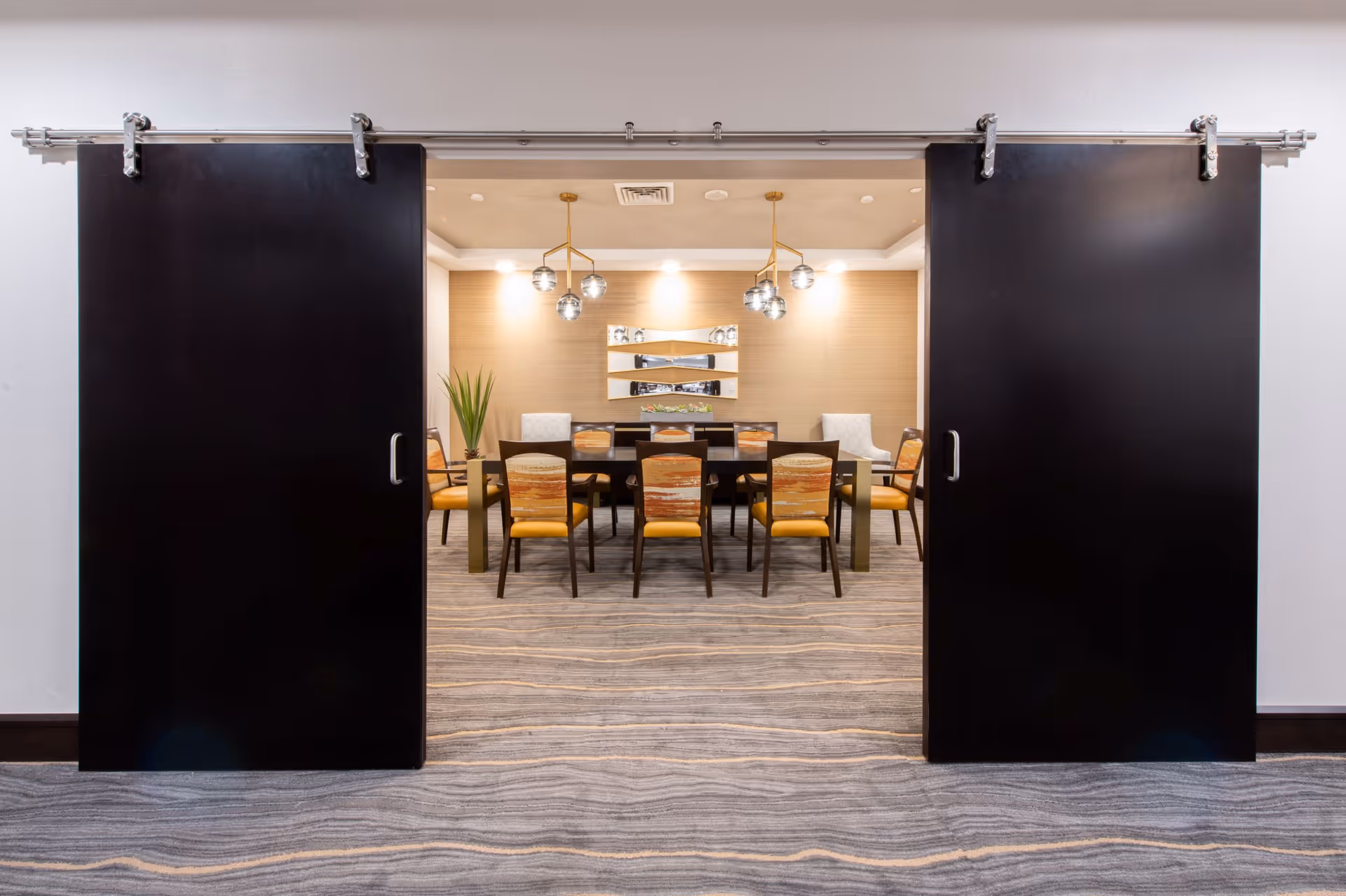 A modern dining room viewed through two large black sliding doors. The room features a long dining table surrounded by chairs with yellow and patterned upholstery. The walls are light-colored with a decorative mirror and two pendant light fixtures hanging from the ceiling. A potted plant is placed in the corner.