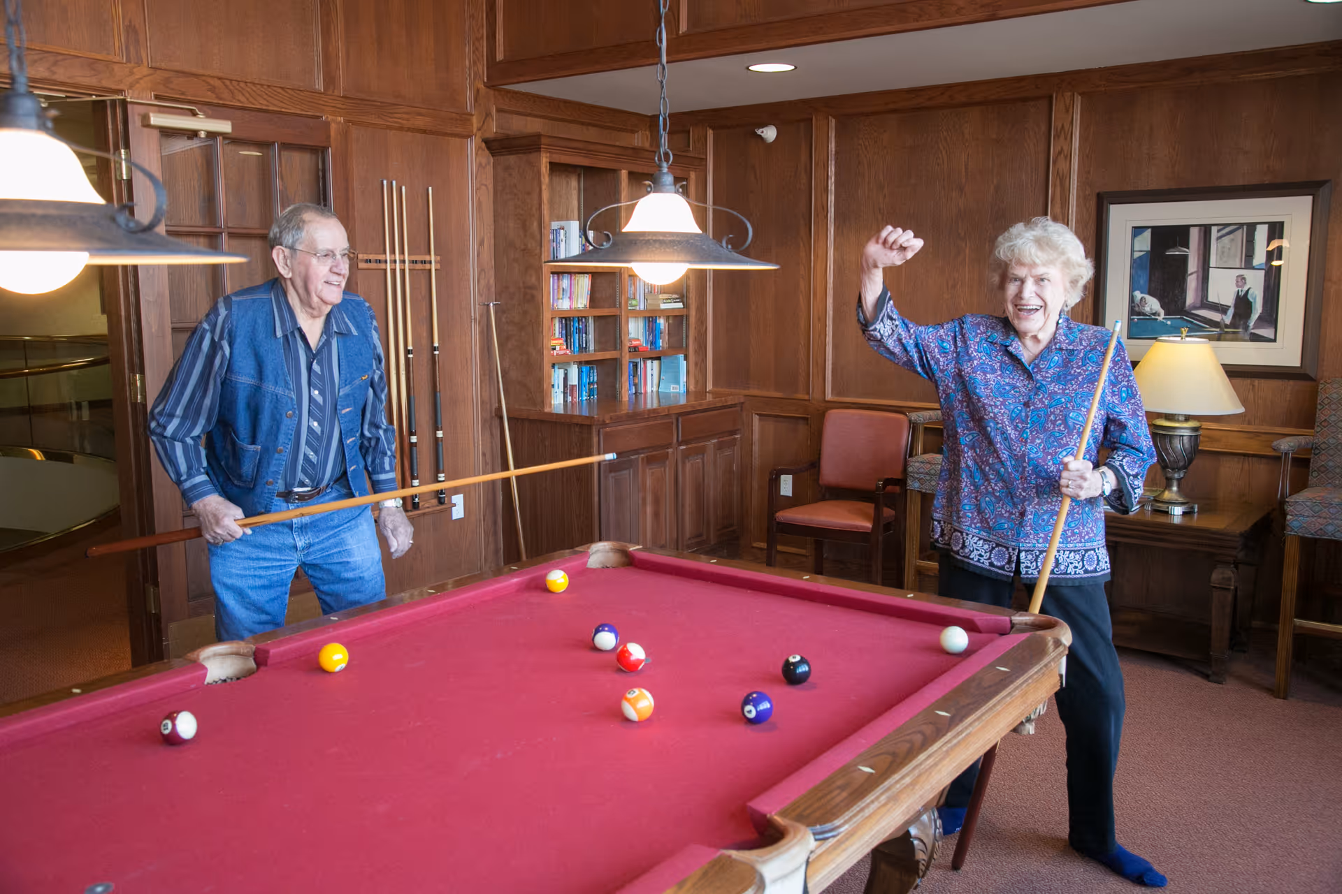 An elderly man and woman playing pool in a wood-paneled room with a red pool table. The man is holding a pool cue and smiling, while the woman is raising her fist in celebration and holding a pool cue. The room has bookshelves, chairs, a lamp, and a framed picture on the wall.