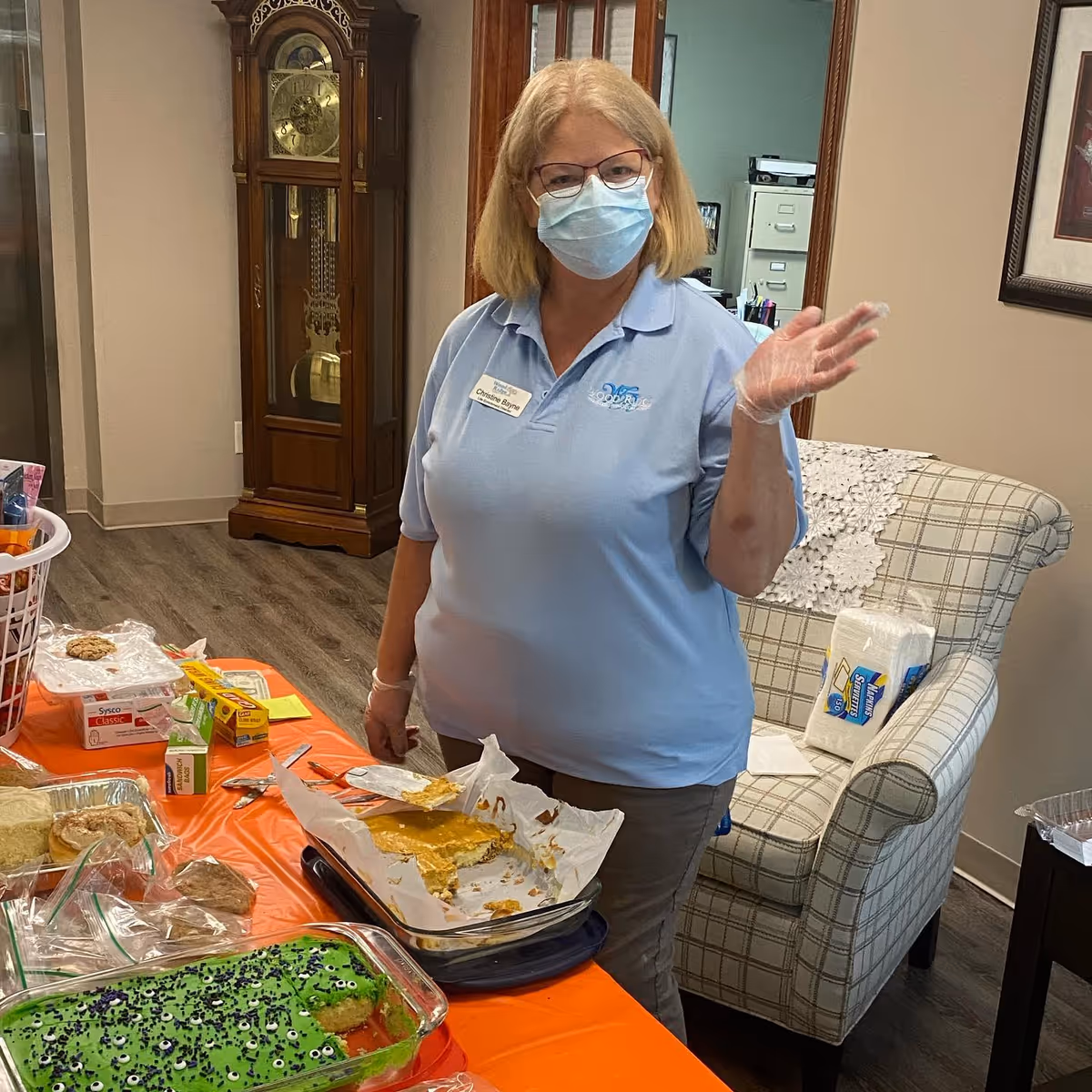 A woman wearing a light blue Wood Ridge Assisted Living polo shirt, a face mask, glasses, and a plastic glove is standing behind a table with various food items and baked goods. Behind her is a plaid armchair with a pack of napkins on it, a wooden grandfather clock, and office filing cabinets.