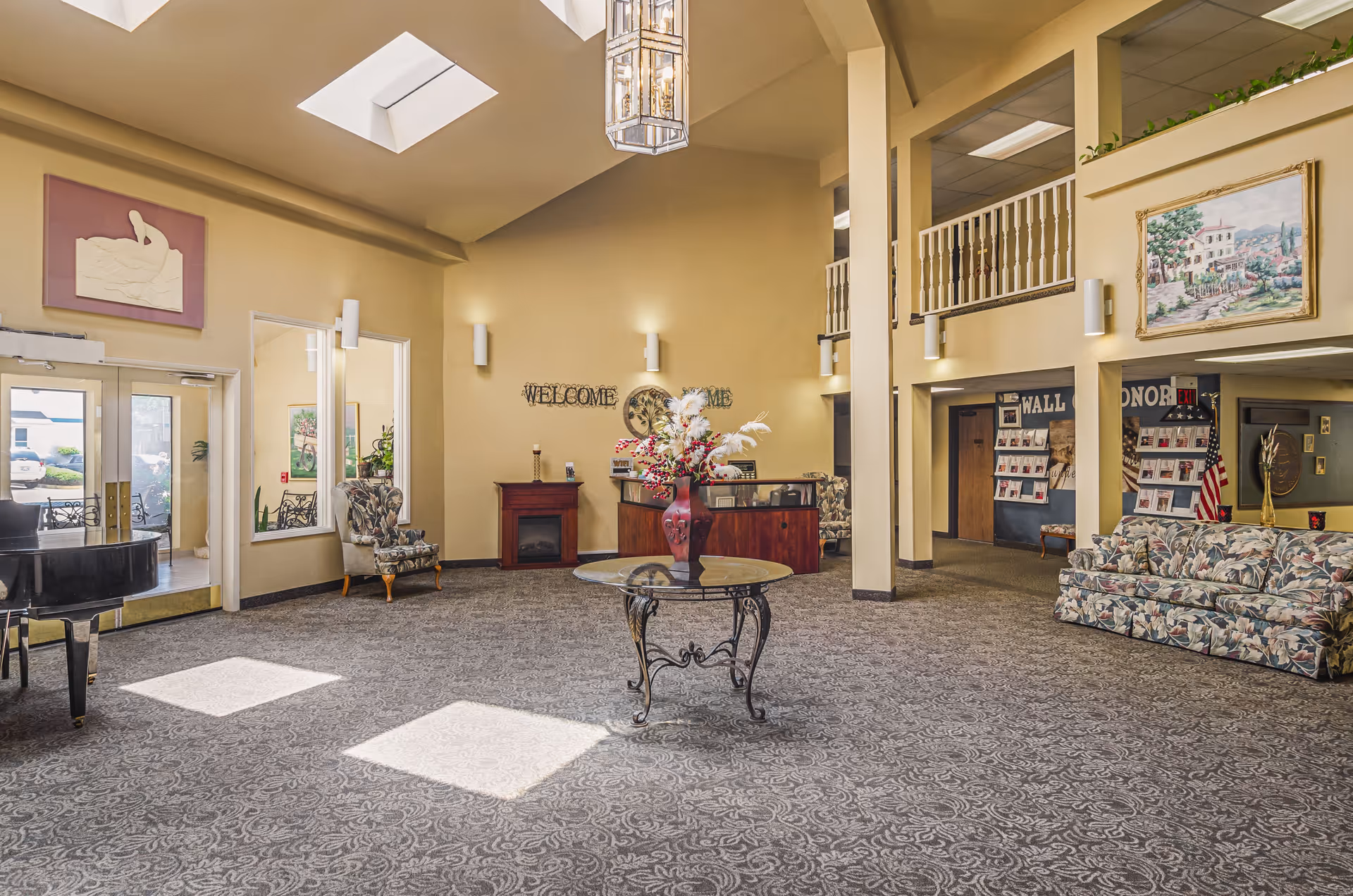 Spacious senior living facility lobby with high ceilings and skylights. The room features a round glass table with a decorative vase of flowers in the center, floral patterned couches and armchairs, a piano near the entrance, a small fireplace, and a reception desk. The walls are decorated with paintings and a 'WELCOME HOME' sign. There is a balcony railing on the upper level and a wall of honor display in the background.