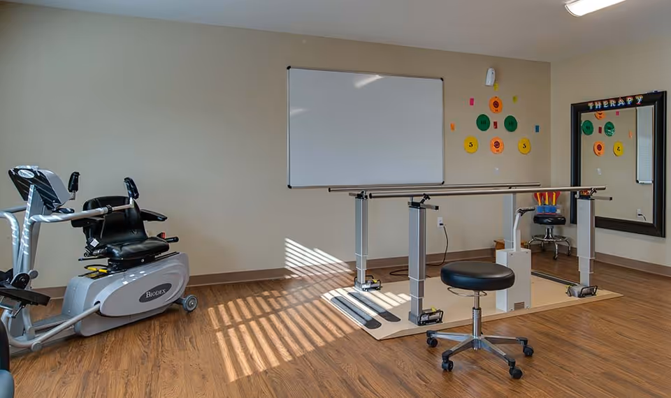 A therapy room with exercise equipment including a recumbent bike and parallel bars for walking practice. There is a black rolling stool, a large mirror on the wall with the word 'THERAPY' above it, a whiteboard, and colorful circular target stickers on the wall. The floor is wood and the room is well-lit with natural light.