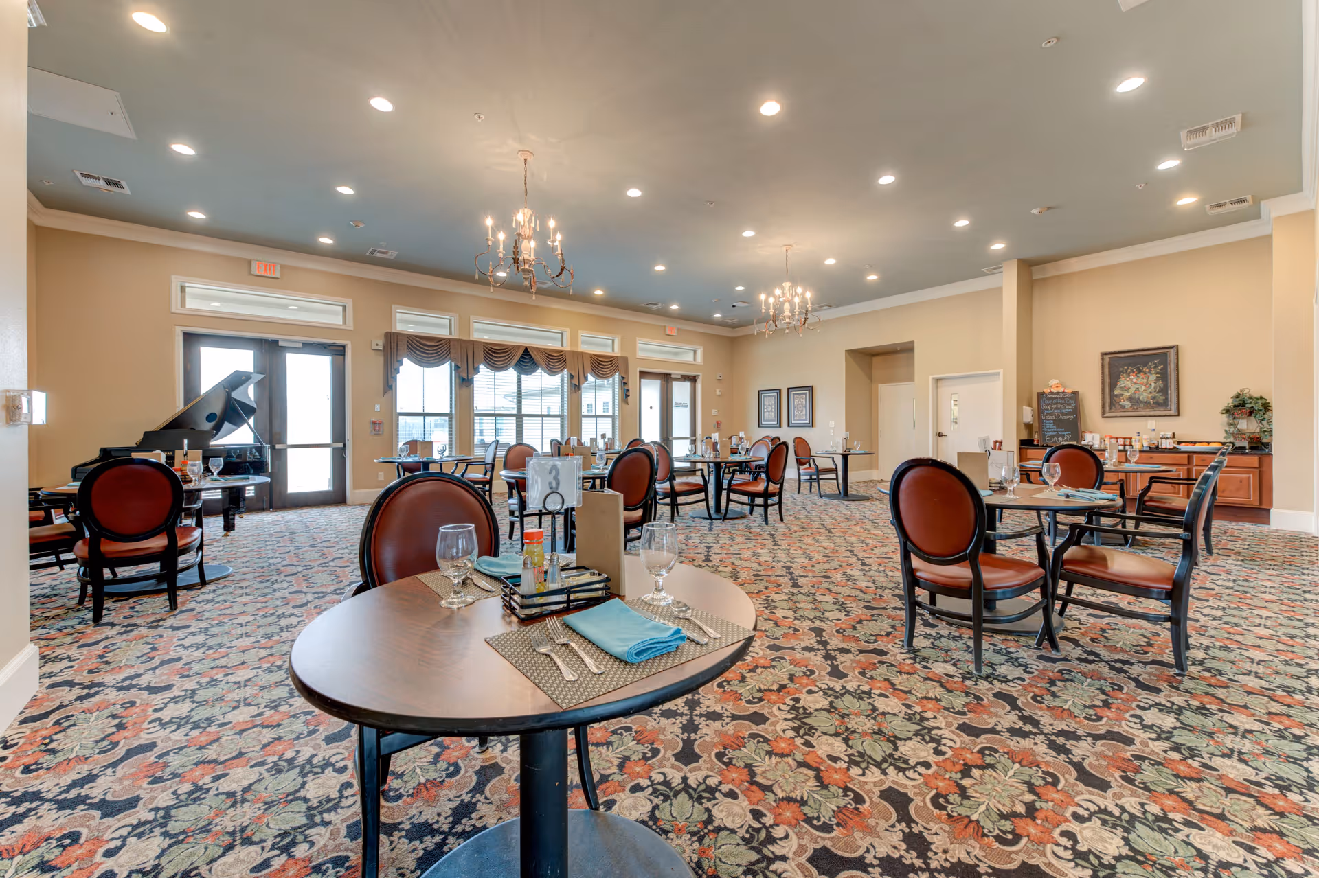 A spacious dining room in a senior living facility with round tables set with placemats, napkins, utensils, and glasses. The room features patterned carpet, beige walls, multiple chandeliers, large windows with valances, and a grand piano in the corner.