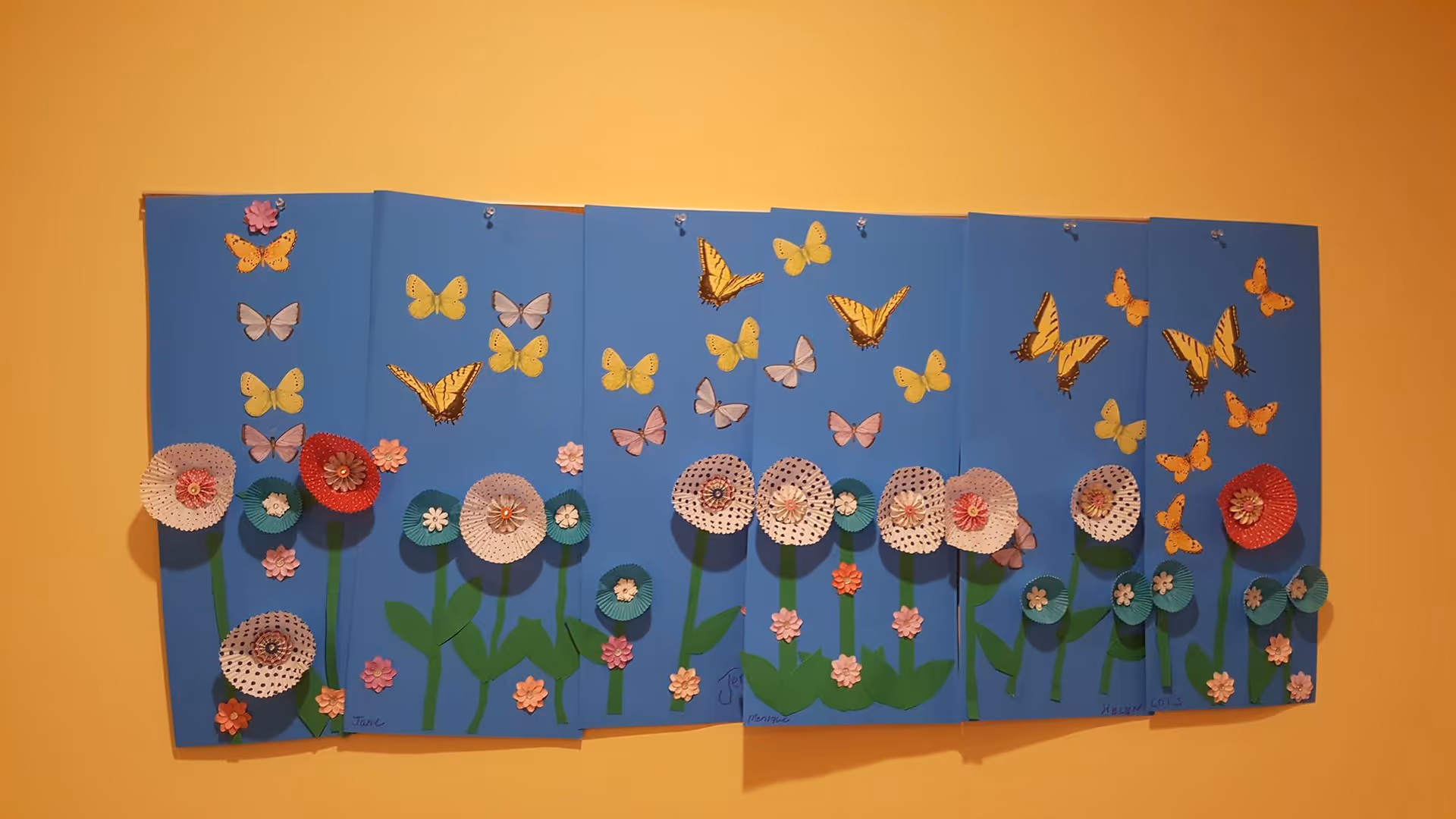 Colorful paper flower and butterfly collage art panels mounted on a yellow wall.